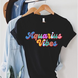 Op de afbeelding: Zwart T-shirt met een kleurrijke retro-lettertypegrafiek die "Aquarius Vibes" zegt.