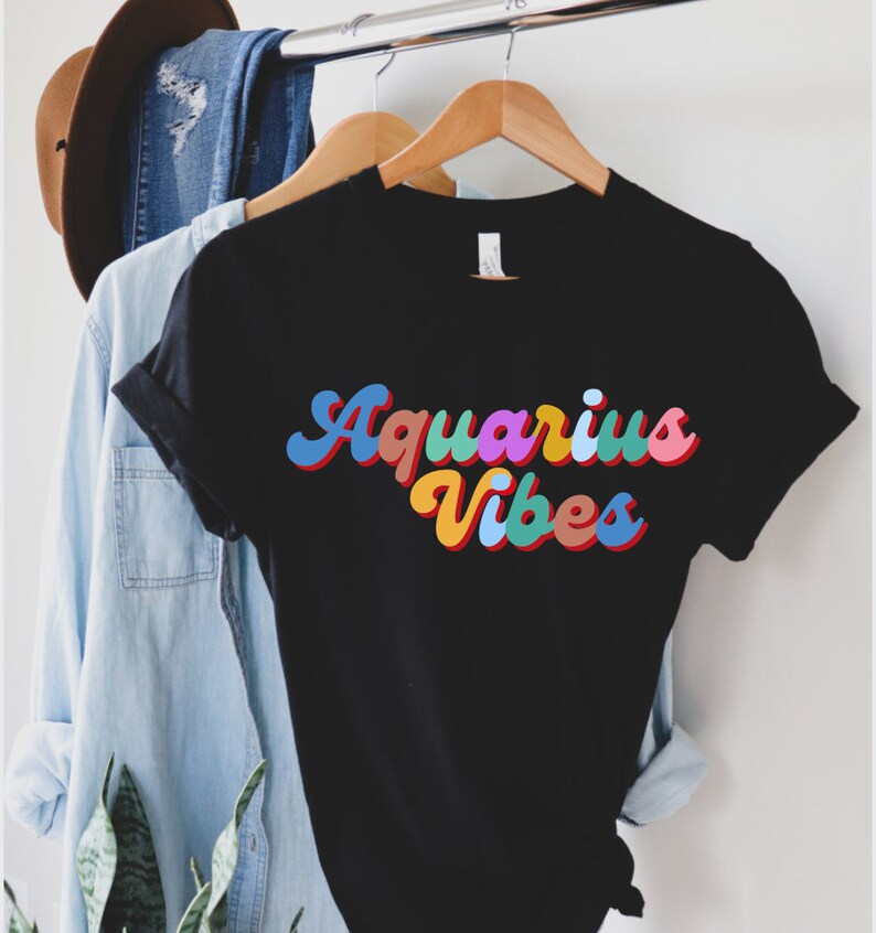 Op de afbeelding: Zwart T-shirt met een kleurrijke retro-lettertypegrafiek die "Aquarius Vibes" zegt.