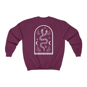 May include: A maroon sweatshirt with a white graphic on the back. The graphic features two intertwined snakes, crescent moons, stars, and the text "FIND YOUR OWN PATH" inside an arched frame. The sweatshirt is a solid color.