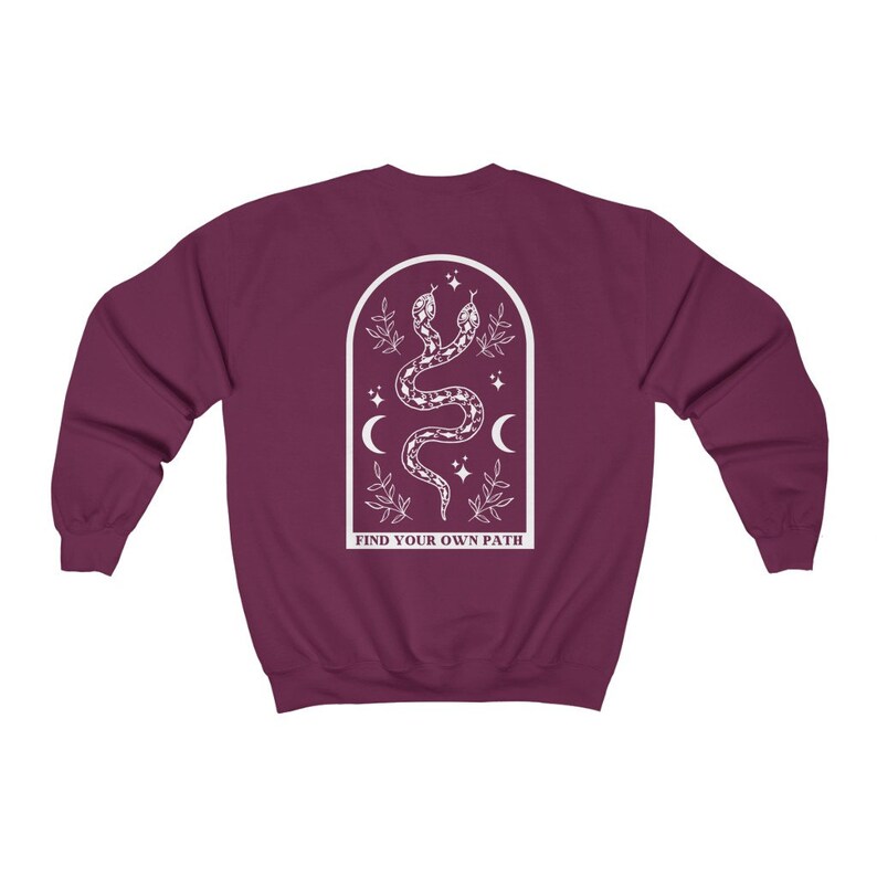 May include: A maroon sweatshirt with a white graphic on the back. The graphic features two intertwined snakes, crescent moons, stars, and the text "FIND YOUR OWN PATH" inside an arched frame. The sweatshirt is a solid color.