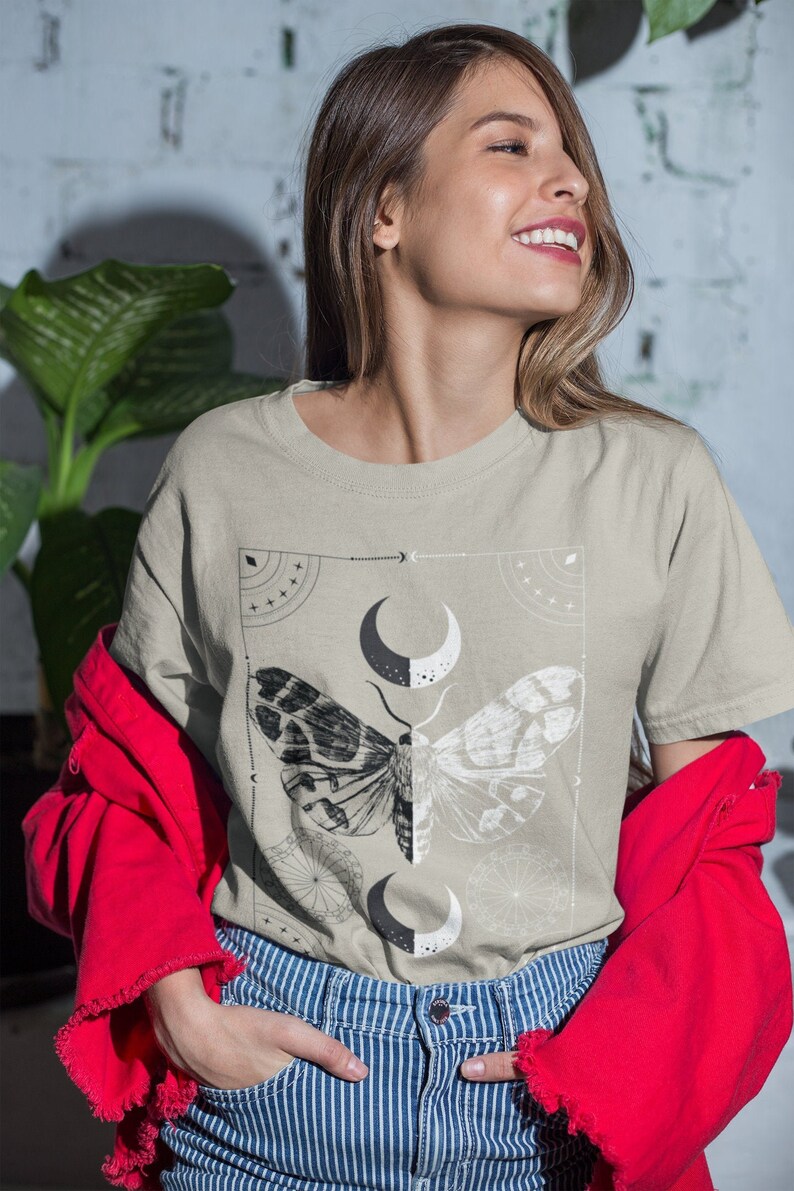 K&ouml;nnte beinhalten: Ein hellbeiges T-Shirt mit einem grafischen Druck eines Nachtfalters und eines Halbmondes. Der Nachtfalter ist schwarzwei&szlig; und hat komplizierte Details. Der Halbmond ist schwarzwei&szlig; und von einem geometrischen Muster umgeben.
