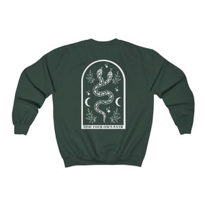 May include: Dark green sweatshirt with a white graphic of two intertwined snakes, crescent moons, stars, and leaves inside an arched frame. The text "FIND YOUR OWN PATH" is below the frame.