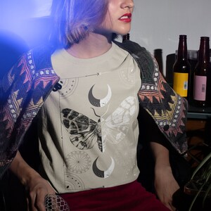 K&ouml;nnte beinhalten: Eine Frau tr&auml;gt ein beiges T-Shirt mit einem schwarzen und wei&szlig;en Motiv aus einem Falter und einem Mond. Das Shirt hat einen Rundhalsausschnitt und kurze &Auml;rmel. Die Frau tr&auml;gt einen roten Rock und eine gemusterte Strickjacke.