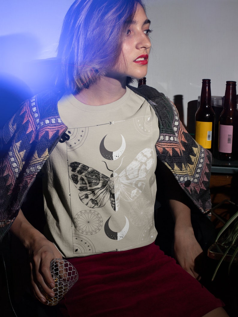 K&ouml;nnte beinhalten: Eine Frau tr&auml;gt ein beiges T-Shirt mit einem schwarzen und wei&szlig;en Motiv aus einem Falter und einem Mond. Das Shirt hat einen Rundhalsausschnitt und kurze &Auml;rmel. Die Frau tr&auml;gt einen roten Rock und eine gemusterte Strickjacke.