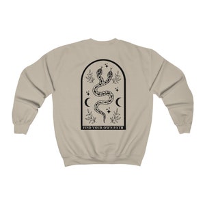 May include: A tan sweatshirt with a black graphic on the back. The graphic features two intertwined snakes within an arched frame, along with moons, stars, and the text "FIND YOUR OWN PATH".