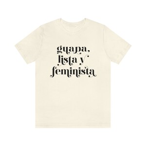 May include: Cream-colored t-shirt with the words "guapa, lista y feminista" in black script. The text is centered on the front of the shirt. The shirt has short sleeves and a crew neck.