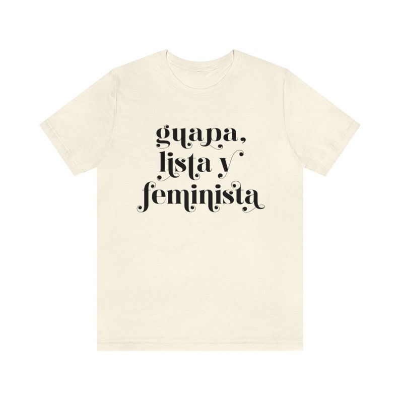 May include: Cream-colored t-shirt with the words "guapa, lista y feminista" in black script. The text is centered on the front of the shirt. The shirt has short sleeves and a crew neck.
