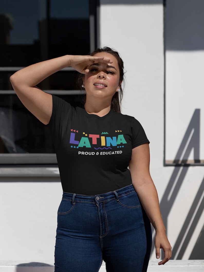 May include: Black t-shirt with the word "LATINA" in colorful geometric letters and the phrase "PROUD & EDUCATED" in white. The model is wearing blue jeans and is shielding her eyes from the sun.