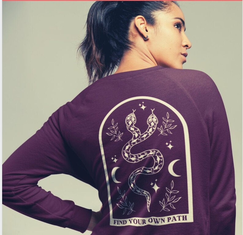 May include: A maroon long-sleeve sweatshirt with a white graphic of two snakes, stars, and crescent moons inside an arch. The text "FIND YOUR OWN PATH" is at the bottom of the graphic.