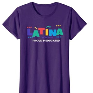 May include: A purple t-shirt with the word "LATINA" in colorful block letters. Below the word is the phrase "PROUD & EDUCATED" in white text. The shirt has short sleeves and a crew neck.