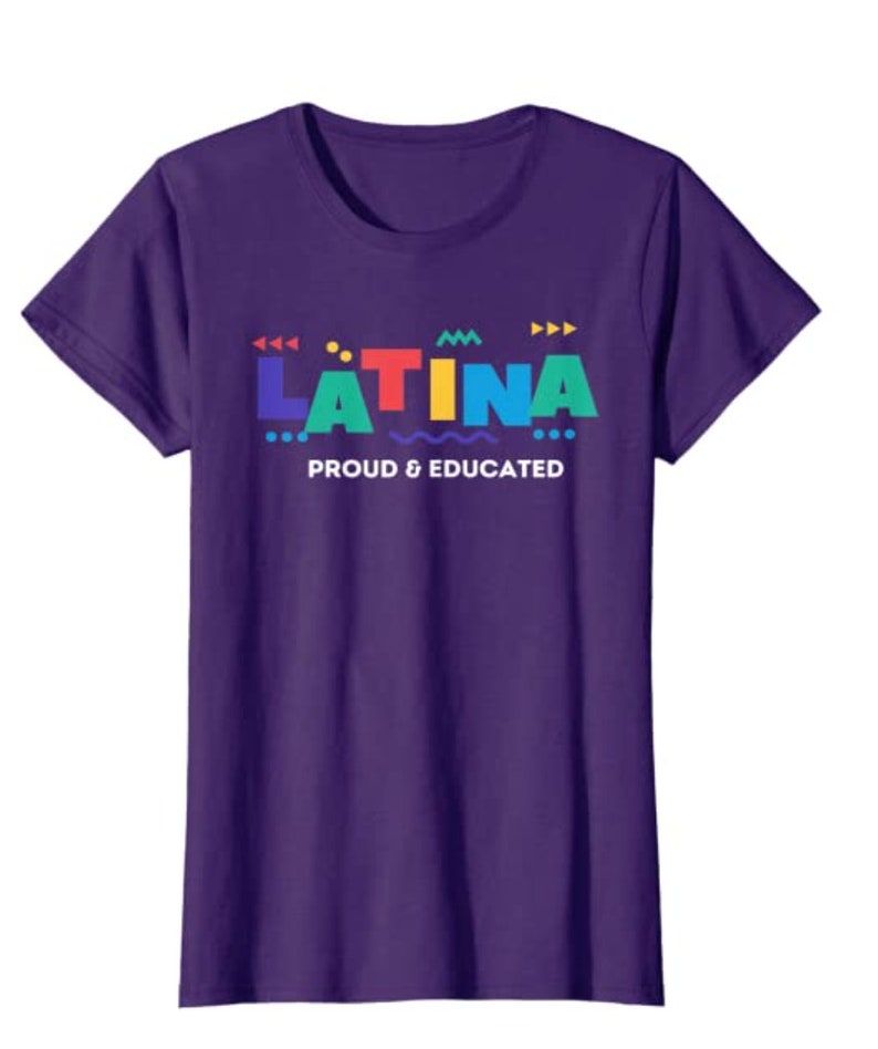May include: A purple t-shirt with the word "LATINA" in colorful block letters. Below the word is the phrase "PROUD & EDUCATED" in white text. The shirt has short sleeves and a crew neck.