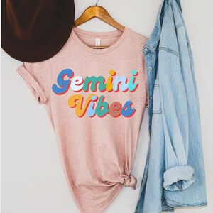 Op de afbeelding: Een roze T-shirt met de tekst "Gemini Vibes" in kleurrijke retro letters.
