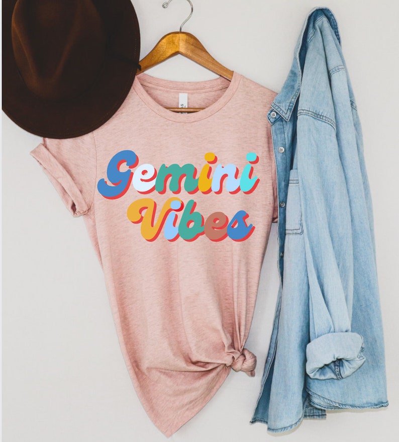 Op de afbeelding: Een roze T-shirt met de tekst "Gemini Vibes" in kleurrijke retro letters.