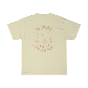 Pode incluir: T-shirt creme com um gr&aacute;fico circular nas costas. O gr&aacute;fico apresenta uma cena de montanha com &aacute;rvores, estrelas e o texto "THE UNIVERSE HAS YOUR BACK" em cor ferrugem. A camisa tem mangas curtas e gola redonda.
