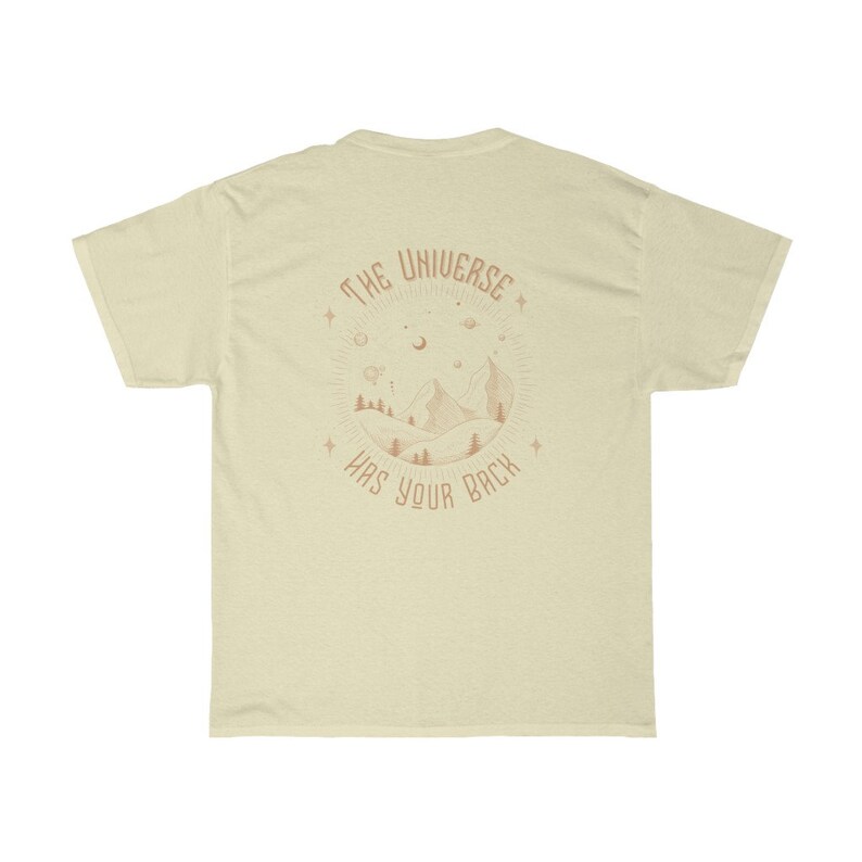 Pode incluir: T-shirt creme com um gr&aacute;fico circular nas costas. O gr&aacute;fico apresenta uma cena de montanha com &aacute;rvores, estrelas e o texto "THE UNIVERSE HAS YOUR BACK" em cor ferrugem. A camisa tem mangas curtas e gola redonda.