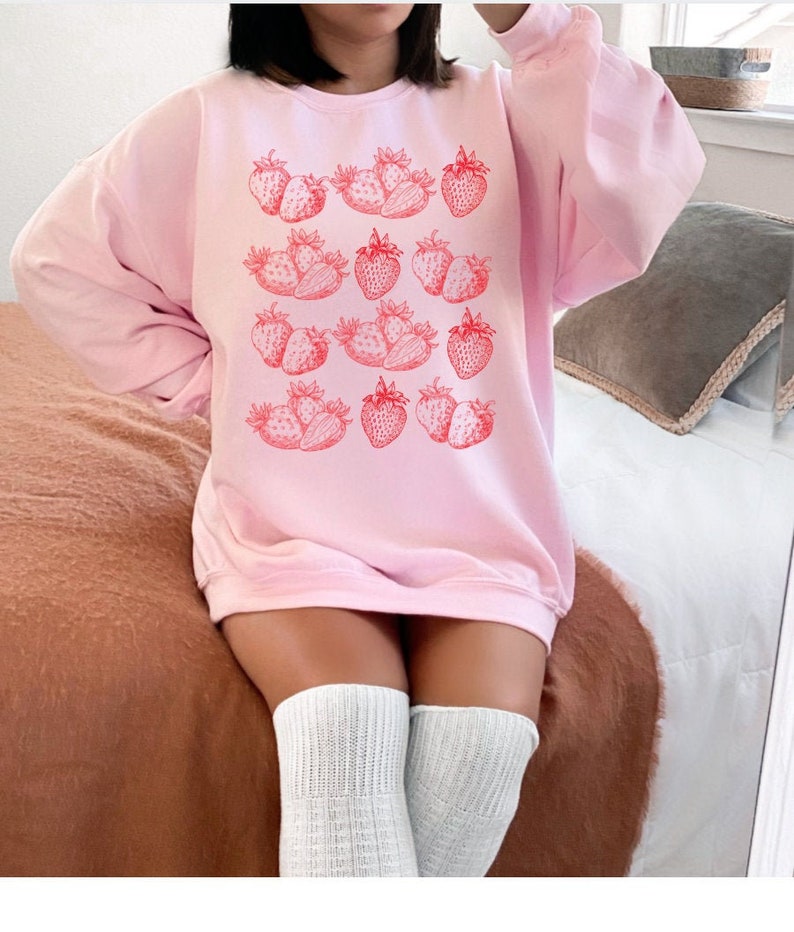 May include: A pink sweatshirt with a graphic print of red strawberries. The sweatshirt has long sleeves and a crew neck.