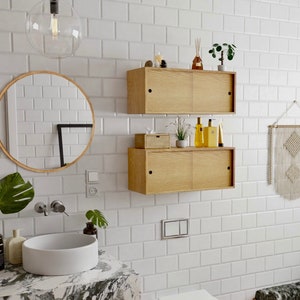 May include: A white bathroom with white subway tile walls, a white porcelain sink and toilet, a wooden cabinet with sliding doors, and a woven wall hanging.