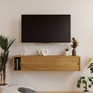 May include: A light wood floating shelf with two sliding doors, a white rabbit figurine, two white ceramic sculptures, and three books titled "The Attic", "Escape", and "2023".