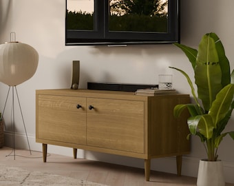 May include: A light wood media console with two doors and black knobs. The console has a flat top with a black sound bar and a glass of water on it. A large flat screen TV is mounted on the wall above the console. A large green plant is in a white pot on the right side of the image. A white lamp with a round shade is on the left side of the image.