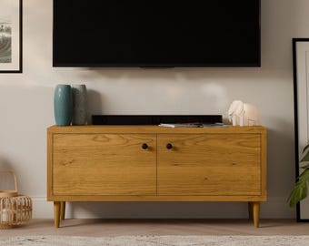 May include: A light wood media console with two doors and black knobs. The console has a light wood finish and sits on four legs. A television is mounted on the wall above the console. There are two vases and a decorative object on top of the console.