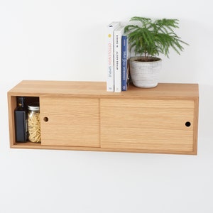 May include: A light oak floating cabinet with sliding doors. The cabinet has a small compartment with a black bottle and a jar of pasta. A small potted plant and books sit on top. The cabinet is mounted on a white wall.