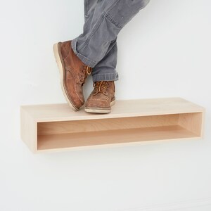 Puede incluir: Una estanter&iacute;a flotante de madera clara con un solo estante abierto. La estanter&iacute;a est&aacute; montada en una pared blanca. Un par de botas de cuero marr&oacute;n descansan sobre la estanter&iacute;a.