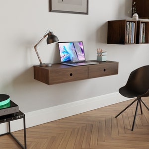 May include: A dark brown floating desk with two drawers, a laptop, a lamp, and a pencil holder on top. The desk is mounted to a white wall above a wood floor.