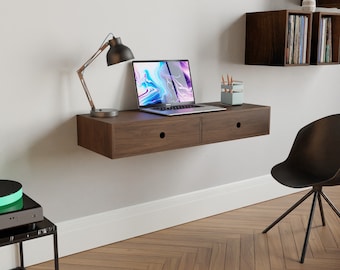 May include: A dark brown floating desk with two drawers, a laptop, a lamp, and a pencil holder on top. The desk is mounted to a white wall above a wood floor.
