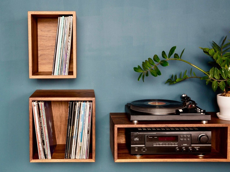 May include: A wooden wall shelf unit with three shelves, two of which are filled with vinyl records. The bottom shelf has a turntable and a stereo receiver on it. A potted plant is on the right side of the image.