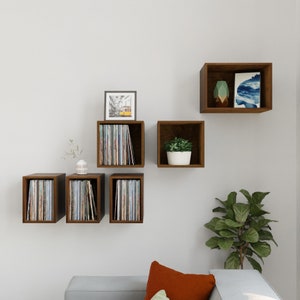 May include: A set of six brown wooden wall shelves, some holding vinyl records, a small potted plant, and a framed picture. The shelves are mounted on a white wall.