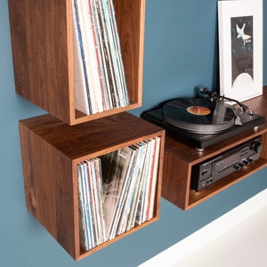 May include: A wall-mounted record player shelf unit with two square compartments and a long shelf. The unit is made of dark wood and has a record player on the top shelf. There are vinyl records in each compartment.
