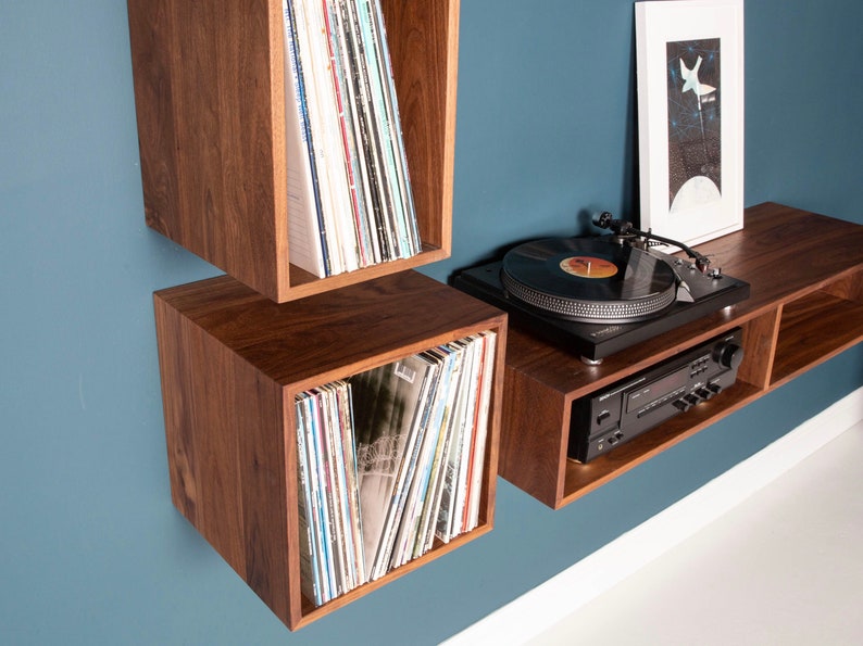 May include: A wall-mounted record player shelf unit with two square compartments and a long shelf. The unit is made of dark wood and has a record player on the top shelf. There are vinyl records in each compartment.