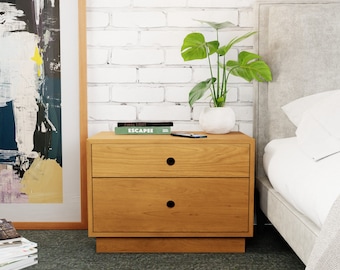 May include: A light wood two-drawer nightstand with a white brick wall and a potted plant in the background. The nightstand has two drawers with black knobs and a small shelf underneath. The nightstand is next to a bed with a white headboard and white bedding.