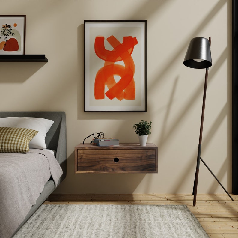 May include: A wooden floating nightstand with a single drawer, a framed abstract painting with orange brushstrokes, a small potted plant, and a floor lamp with a black shade.