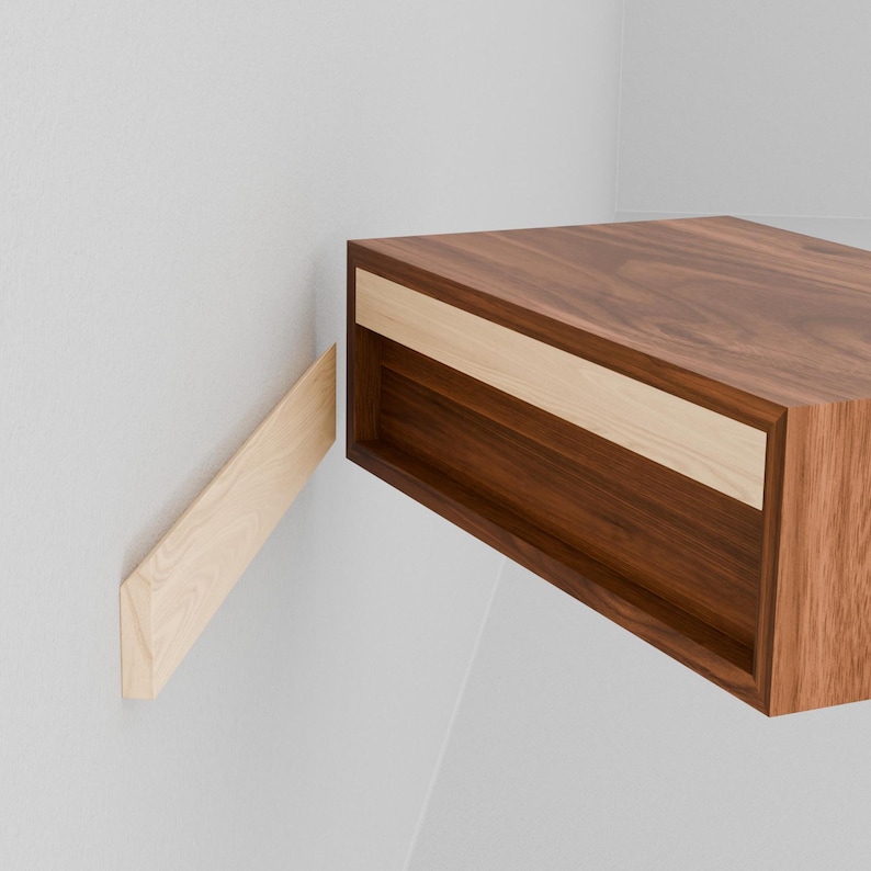 May include: A floating walnut wood nightstand with a drawer. The drawer front is made of a lighter wood. The nightstand is mounted to the wall with a wooden bracket.