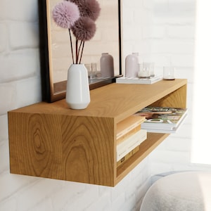 May include: A light wood floating shelf with a white vase and pink flowers. The shelf has a built-in cubby for storing books and magazines. The shelf is mounted on a white brick wall.
