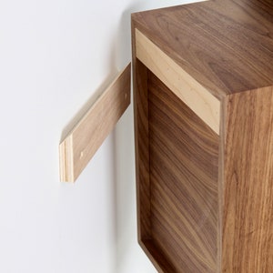 May include: A wooden wall shelf with a light wood finish is being installed on a white wall. The shelf is being held in place by a person's hand. The shelf has a simple design and is made of solid wood.
