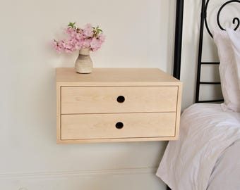 May include: A light wood floating bedside table with two drawers and rounded cutouts for handles. The bedside table is mounted to a white wall and has a vase of pink flowers on top.