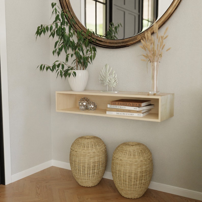 Puede incluir: Una estanter&iacute;a de madera clara con fondo blanco, con una peque&ntilde;a planta en maceta, dos esferas decorativas y una pila de libros con el t&iacute;tulo "The Fashion Book". La estanter&iacute;a est&aacute; montada en una pared blanca, y hay dos cestas tejidas en el suelo debajo.