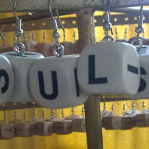 Pode incluir: Um par de brincos feitos de dados brancos com letras pretas que soletram "PUL-I-L" pendurados em ganchos prateados.