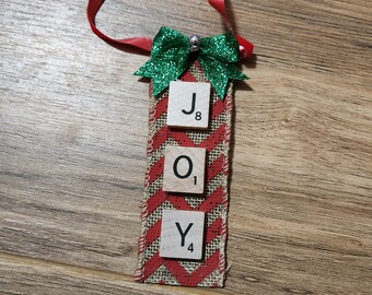 May include: A Christmas ornament made from burlap ribbon with red and white stripes. The ornament features a green glitter bow and three wooden Scrabble tiles spelling out the word "JOY".
