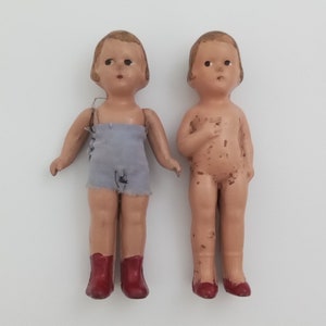 May include: Two vintage porcelain dolls with painted features. One doll is wearing a blue and white outfit and red shoes. The other doll is nude and has brown spots on its body. Both dolls have brown hair.