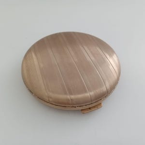 May include: A round, gold-toned compact with a textured, diagonal striped pattern on the lid. The compact has a small clasp and a slightly raised edge. The overall design is simple and elegant.