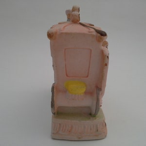 May include: A pink ceramic figurine of a carriage with a yellow seat and a white roof. The carriage is pulled by two horses and has a driver in the front. The figurine is on a white base.