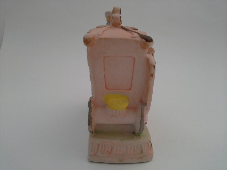 May include: A pink ceramic figurine of a carriage with a yellow seat and a white roof. The carriage is pulled by two horses and has a driver in the front. The figurine is on a white base.