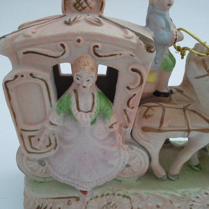 May include: A porcelain figurine of a horse-drawn carriage with a woman and a man riding inside. The carriage is pink with gold accents and the horses are white with brown harnesses. The woman is wearing a green and pink dress and the man is wearing a blue shirt and brown pants.