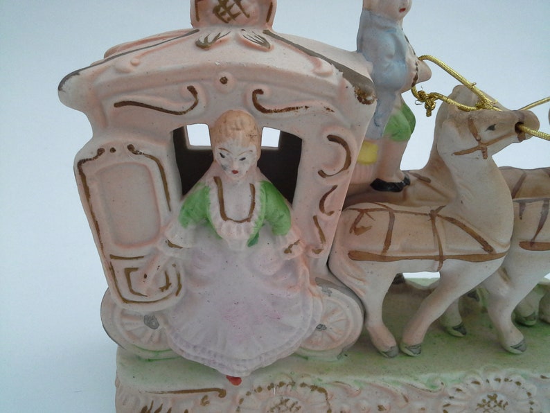 May include: A porcelain figurine of a horse-drawn carriage with a woman and a man riding inside. The carriage is pink with gold accents and the horses are white with brown harnesses. The woman is wearing a green and pink dress and the man is wearing a blue shirt and brown pants.