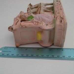 May include: A porcelain figurine of a woman riding in a carriage pulled by two horses. The figurine is painted in pink, white, and gold tones. The carriage has a detailed design and the horses have flowing manes. The figurine is approximately 5 inches tall.
