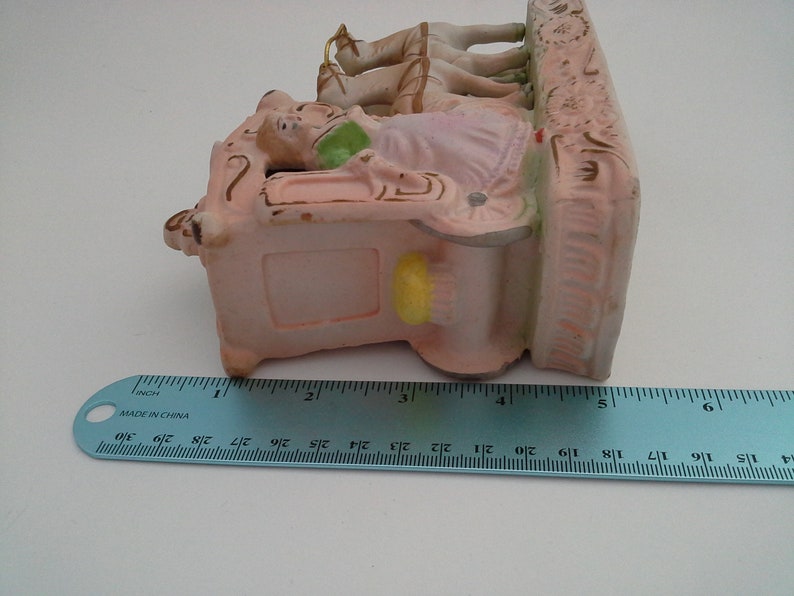 May include: A porcelain figurine of a woman riding in a carriage pulled by two horses. The figurine is painted in pink, white, and gold tones. The carriage has a detailed design and the horses have flowing manes. The figurine is approximately 5 inches tall.