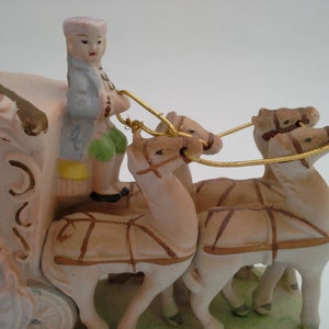May include: A porcelain figurine of a horse-drawn carriage with a driver in a blue and green outfit. The carriage is pink and white with gold accents. The horses are brown and white.